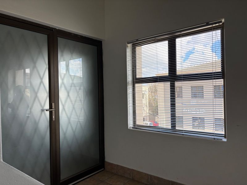 A bright, empty office space featuring tiled flooring, a frosted glass door, and a large window with fitted venetian blinds.