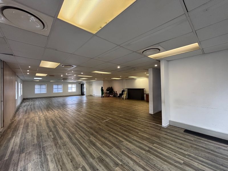 A spacious, open-plan office area featuring laminate flooring, a suspended ceiling with recessed lighting, and large windows providing natural light.