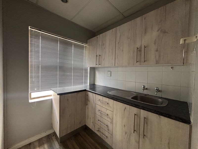 A compact office kitchenette featuring light wood-look cabinetry, a stainless steel sink, dark laminate countertops, and a tiled backsplash.