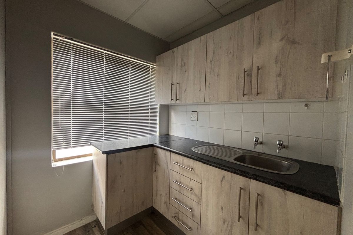 A compact office kitchenette featuring light wood-look cabinetry, a stainless steel sink, dark laminate countertops, and a tiled backsplash.