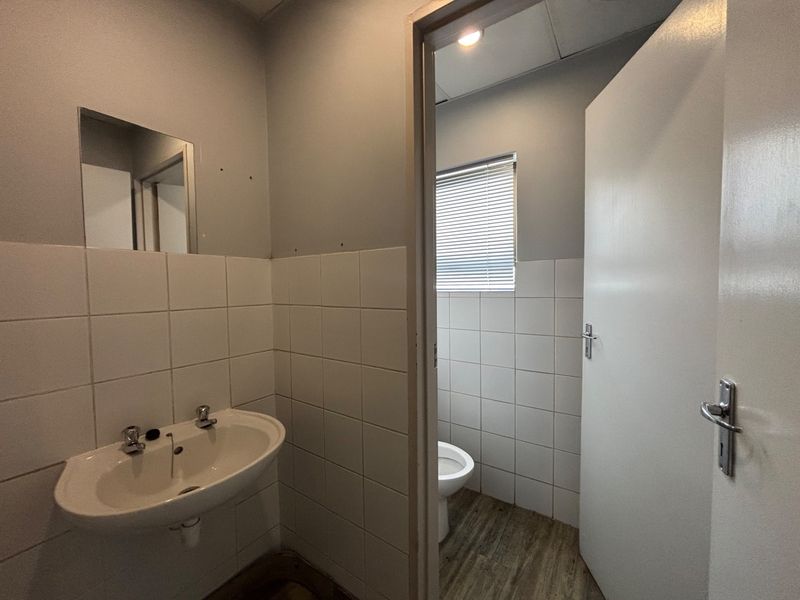 A functional office bathroom featuring a wall-mounted basin and separate toilet cubicle with tiled splashbacks and neutral finishes.