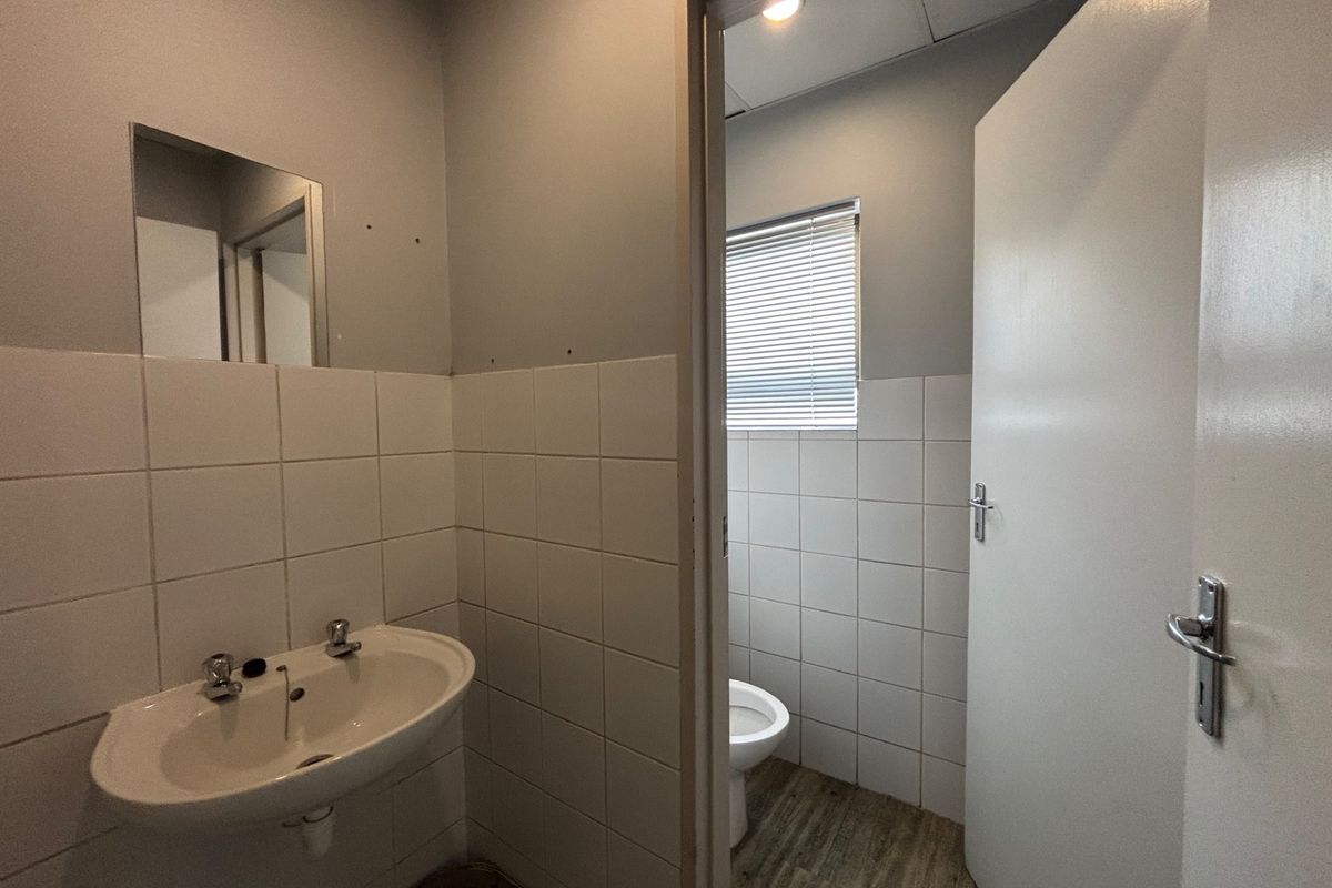 A functional office bathroom featuring a wall-mounted basin and separate toilet cubicle with tiled splashbacks and neutral finishes.
