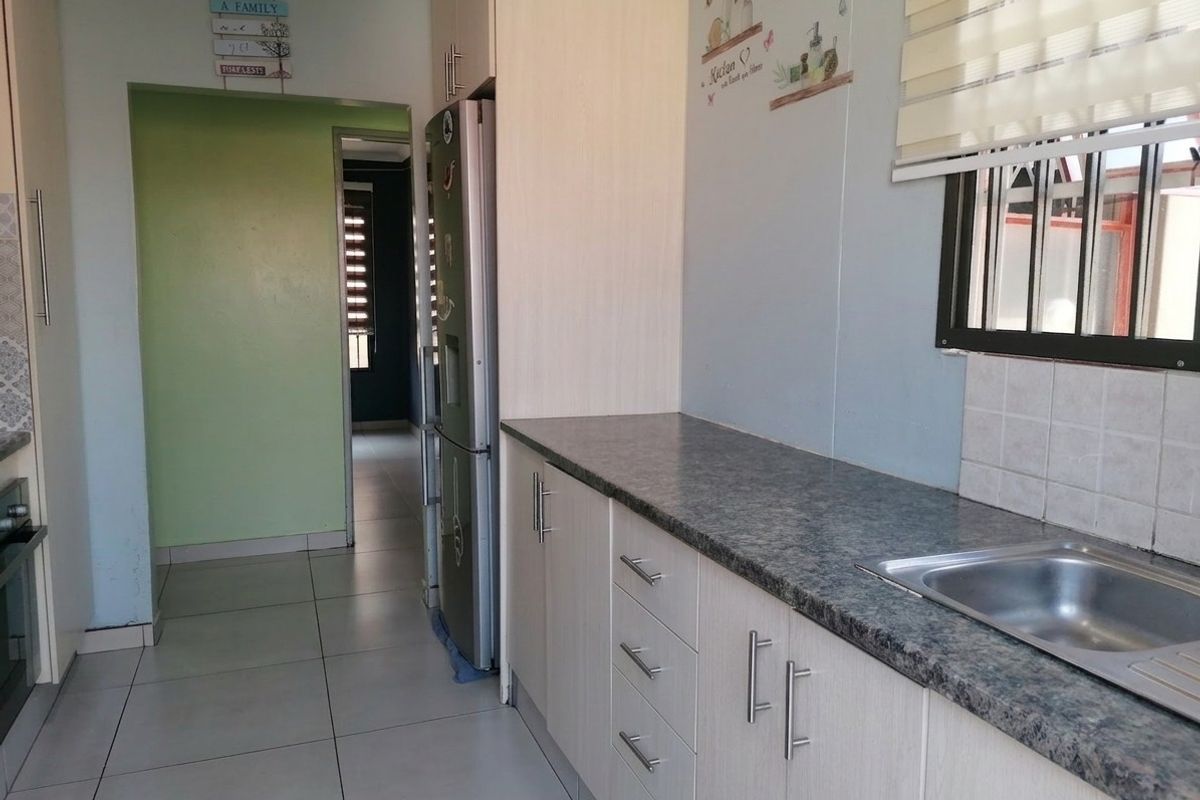 A functional kitchen featuring light-toned cabinetry, durable laminate countertops, a stainless steel sink, and tiled flooring with a view into the adjacent hallway.
