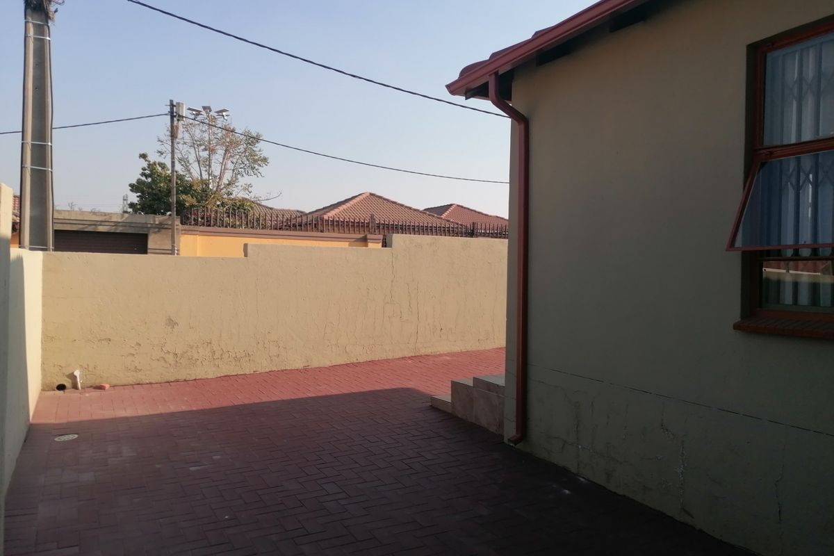 A rear exterior view showing a paved courtyard area, boundary wall, and the side elevation of the house with security window bars.