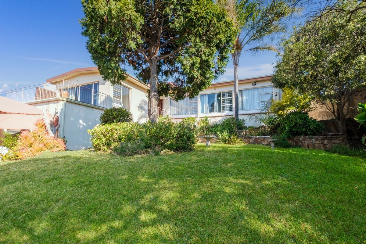 Rear view of the house featuring a raised patio with metal balustrade, overlooking a well-maintained garden with stone retaining walls and mature trees.