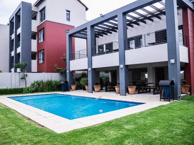 A well-maintained outdoor pool area featuring a paved surround, covered patio with pergola, and landscaped garden within a modern apartment complex.