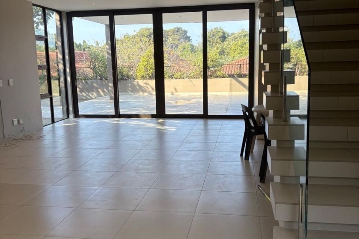 Spacious open-plan living area featuring large sliding glass doors, modern tiled flooring, and a contemporary floating staircase with a glass balustrade.