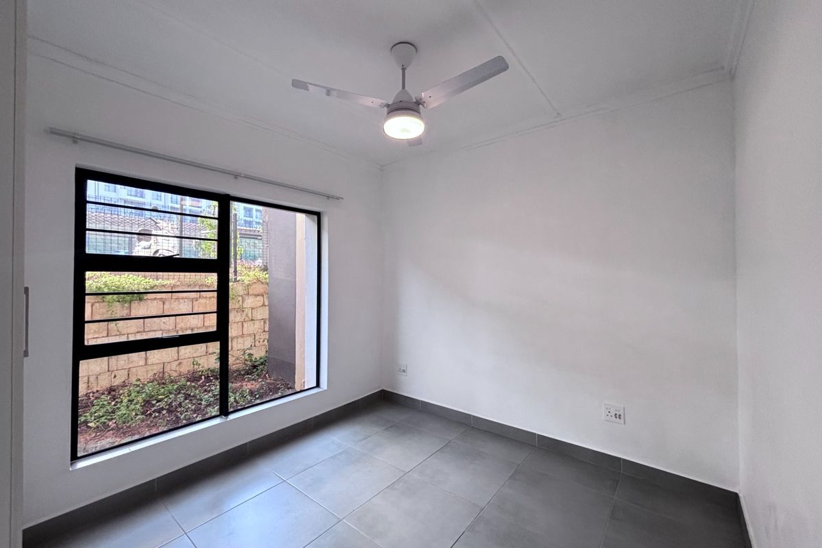 A clean, empty bedroom featuring modern grey floor tiles, a ceiling fan, and a large window with security bars overlooking a garden area.
