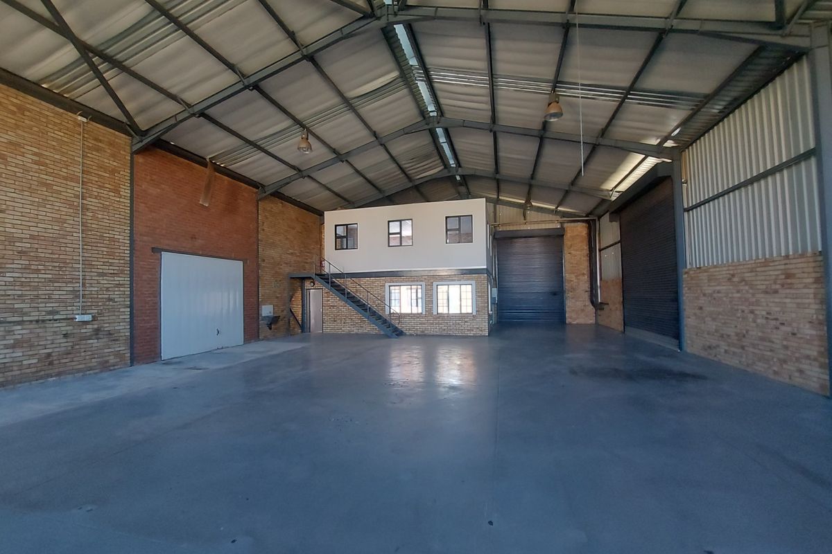 Spacious industrial warehouse featuring high ceilings with exposed steel trusses, concrete flooring, roller shutter access, and an integrated mezzanine office level.