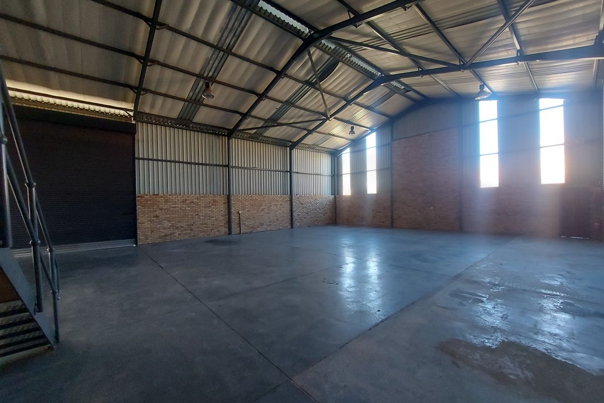 Spacious industrial warehouse unit featuring high ceilings with exposed steel trusses, durable concrete flooring, face brick walls, and a large roller shutter door.