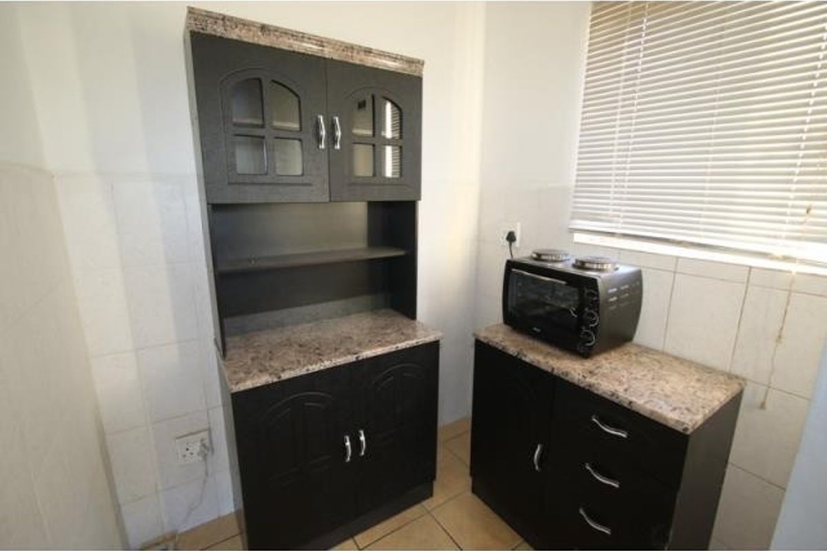 A compact kitchen area featuring dark built-in cabinetry, stone-look laminate countertops, tiled splashbacks, and a window fitted with venetian blinds.