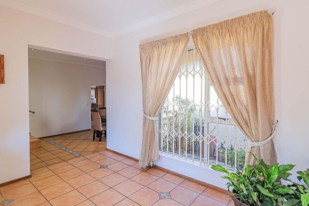 A bright, open-plan living area featuring ceramic floor tiles, coved ceilings, and a window fitted with a security gate.