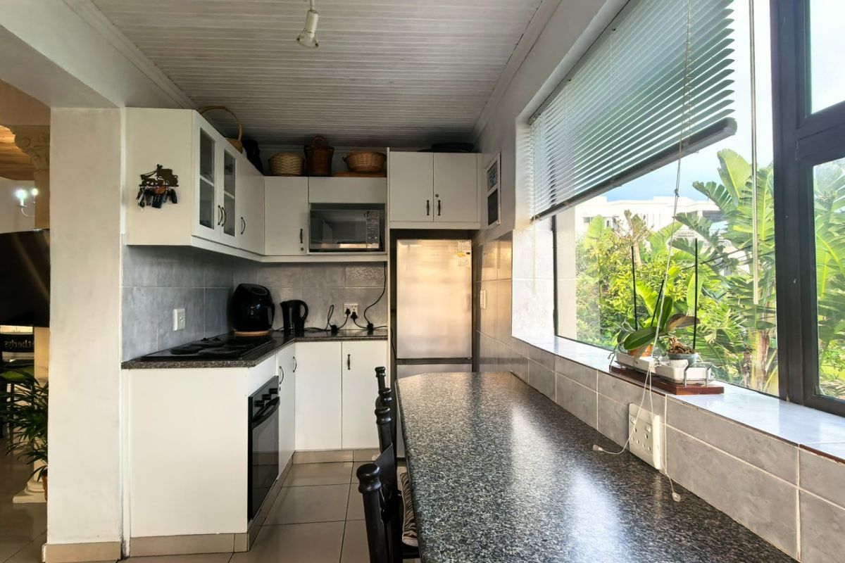 Compact kitchen featuring white cabinetry, granite countertops, built-in oven and hob, and a breakfast bar overlooking the garden through a large window.