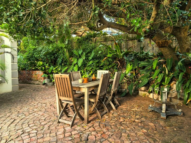 A charming, shaded outdoor patio area featuring cobblestone paving, surrounded by lush mature trees and established garden landscaping.