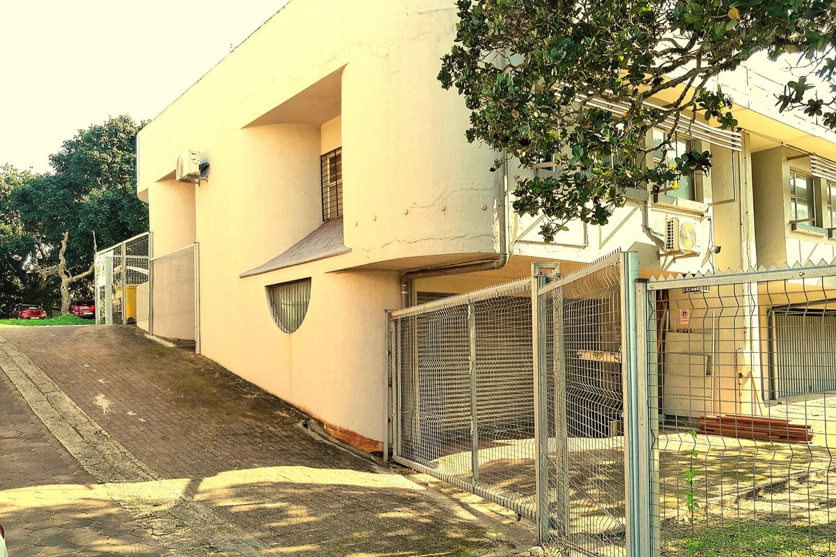 Rear exterior view of a commercial building featuring a paved driveway, security fencing, and weathered concrete facade with visible plaster cracking.