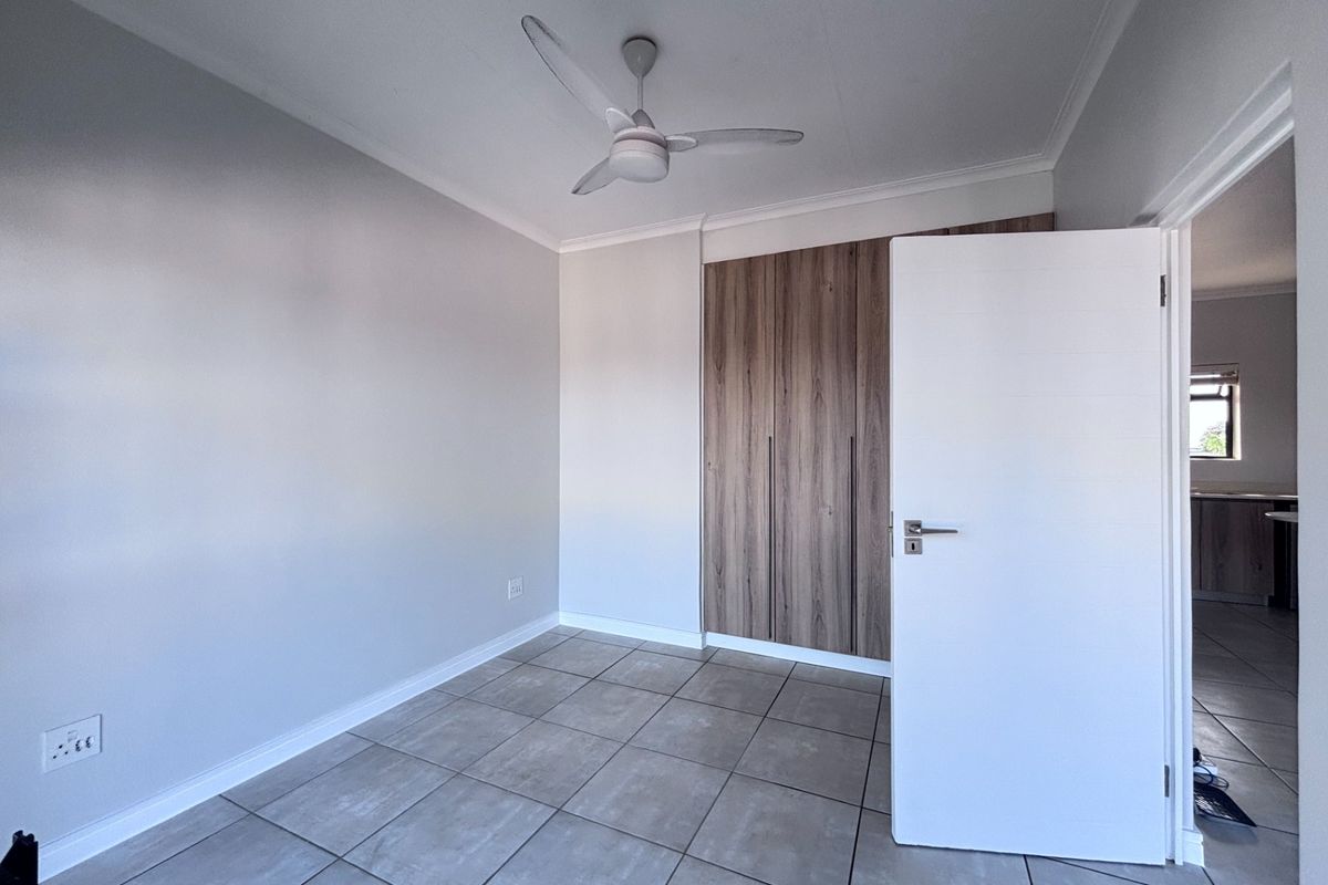 A clean, empty bedroom featuring modern wood-finish built-in cupboards, neutral tiled flooring, and a ceiling fan.