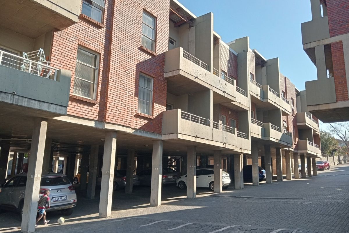 Exterior view of a multi-story apartment complex featuring exposed brickwork, concrete balconies, and a covered ground-level parking area.