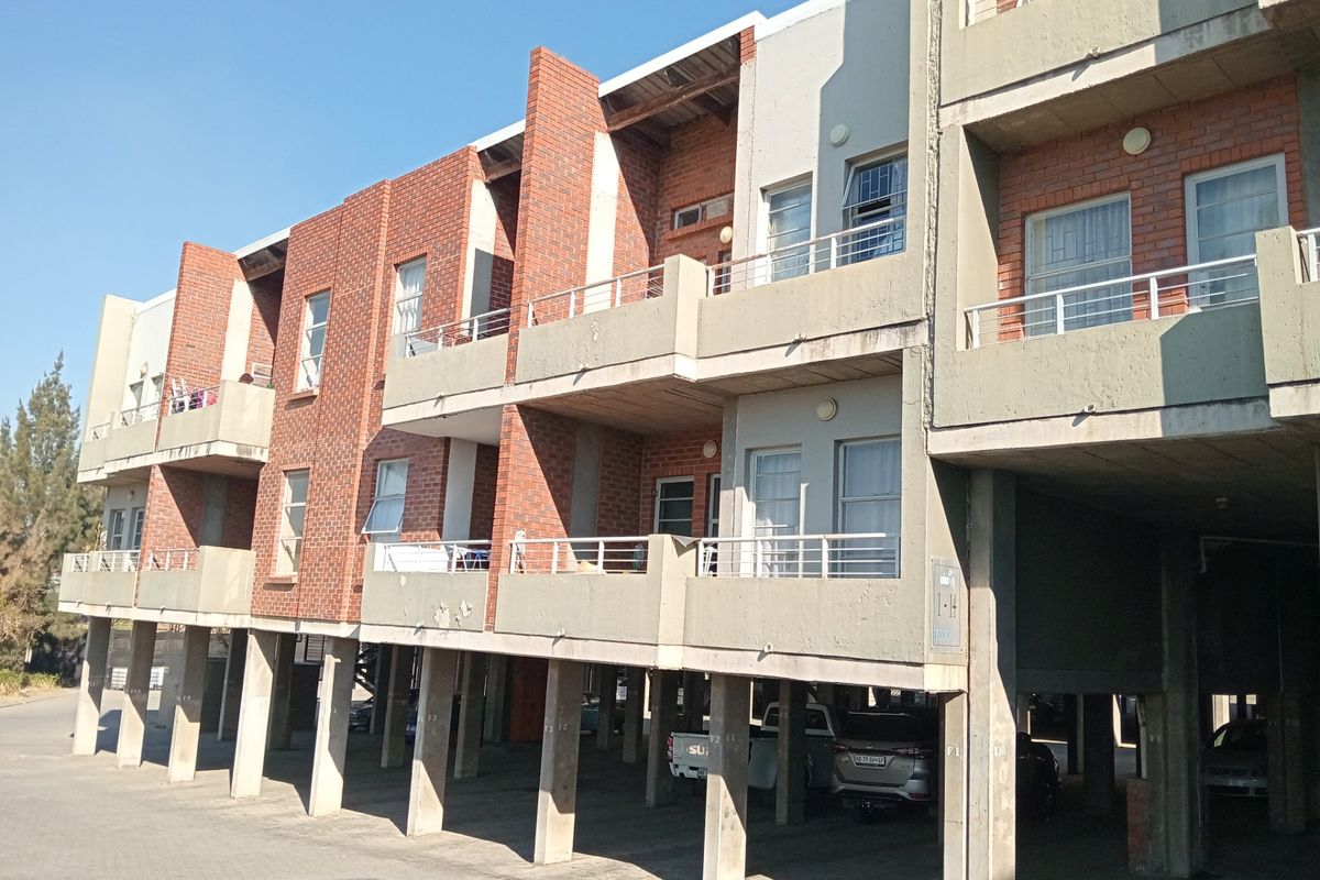 Exterior view of a multi-story apartment building featuring exposed brickwork, concrete balconies with metal railings, and a ground-level covered parking area.