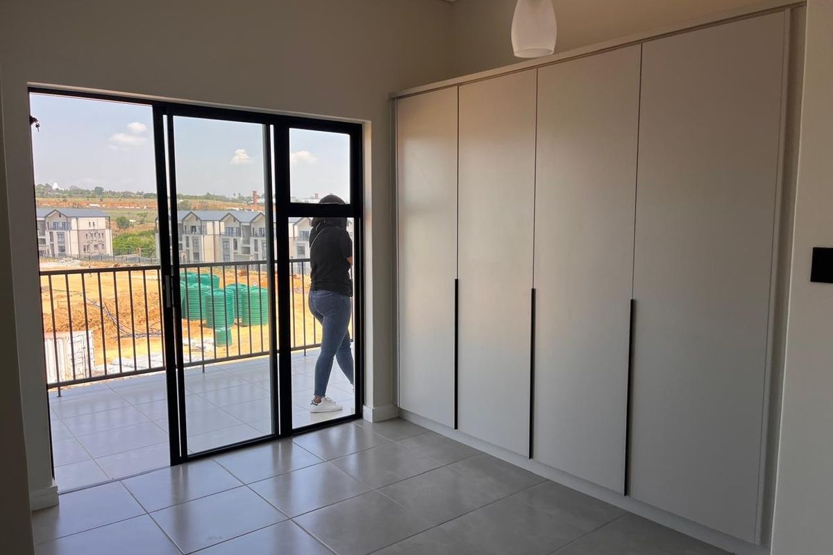A bright, modern bedroom featuring neutral tiled floors, ample built-in cupboard space, and a sliding door leading to a private balcony.