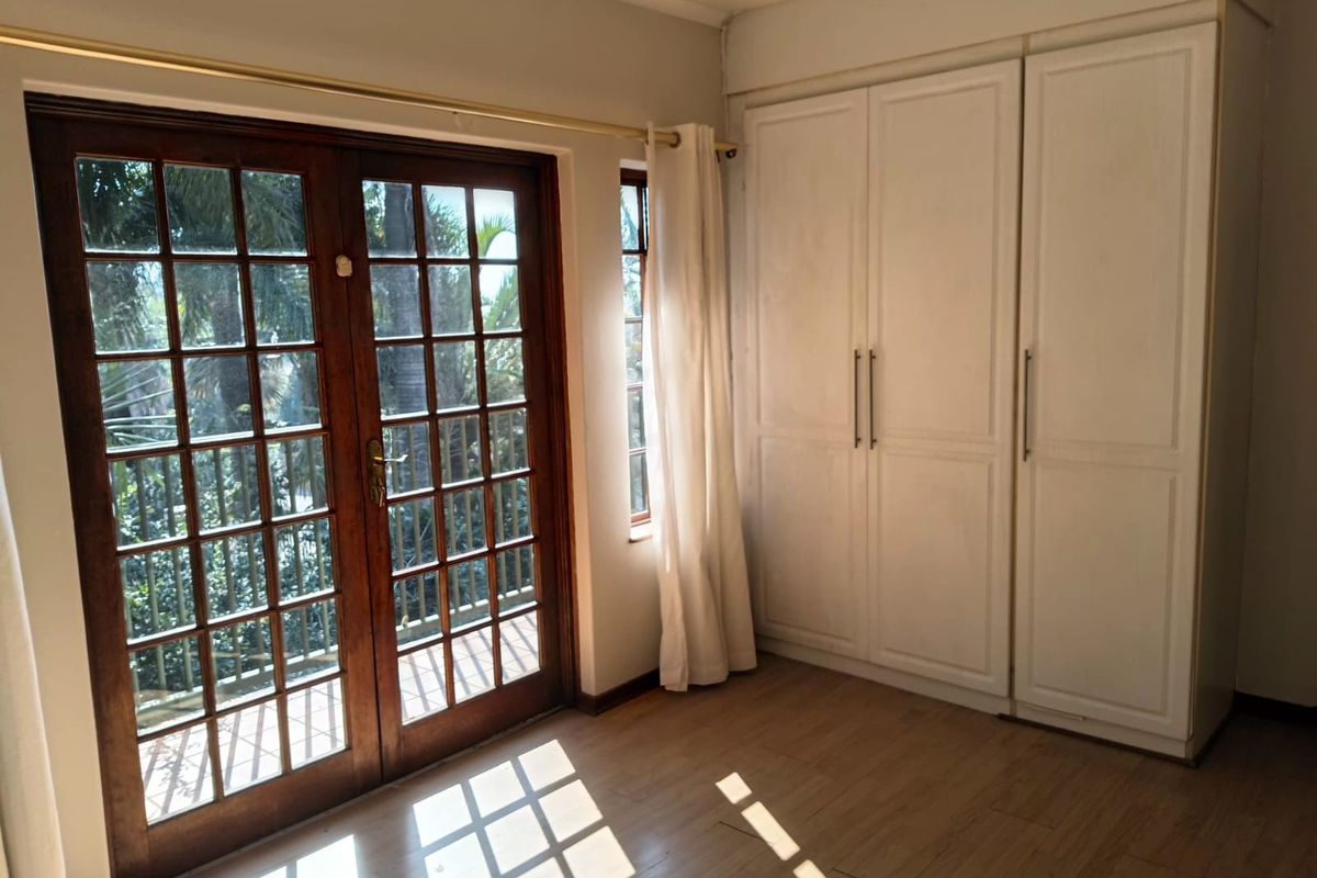 A bright, empty bedroom featuring laminate flooring, built-in cupboards, and wooden french doors opening to an outdoor area.