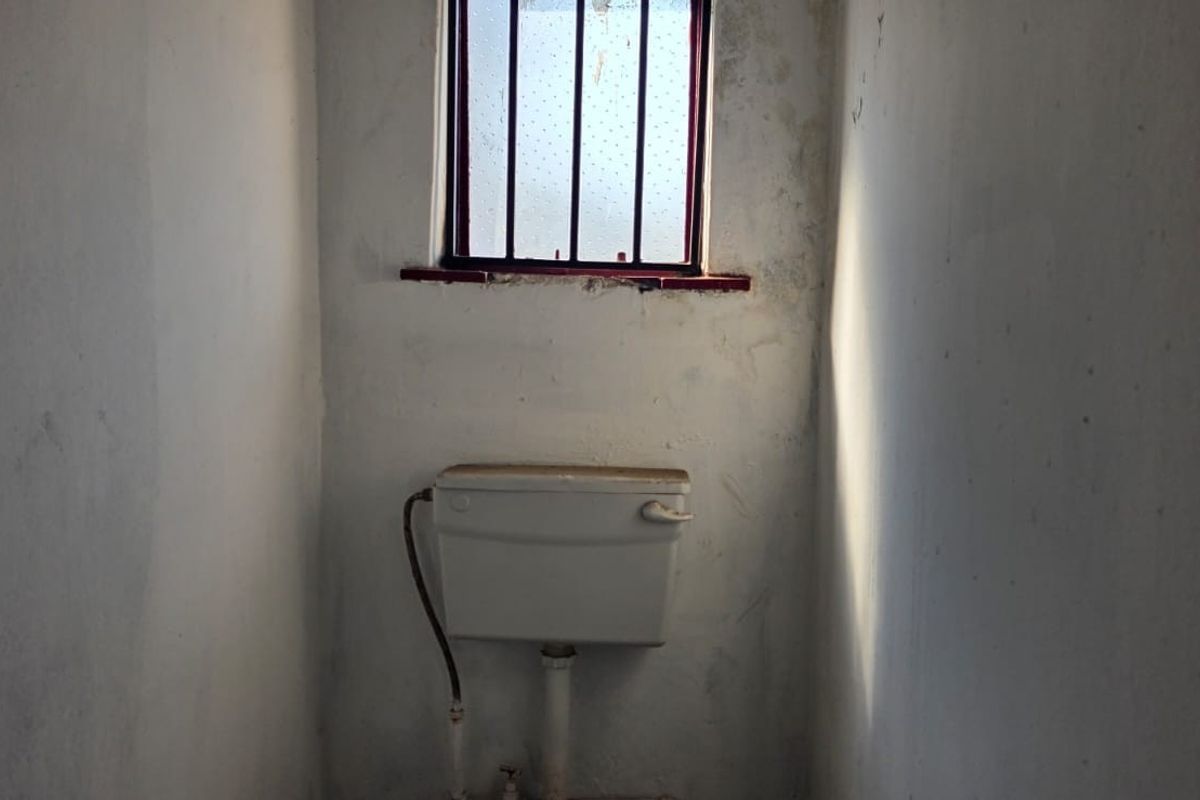 A small, basic bathroom space featuring a wall-mounted toilet cistern and a window with security bars, requiring significant cosmetic refurbishment.