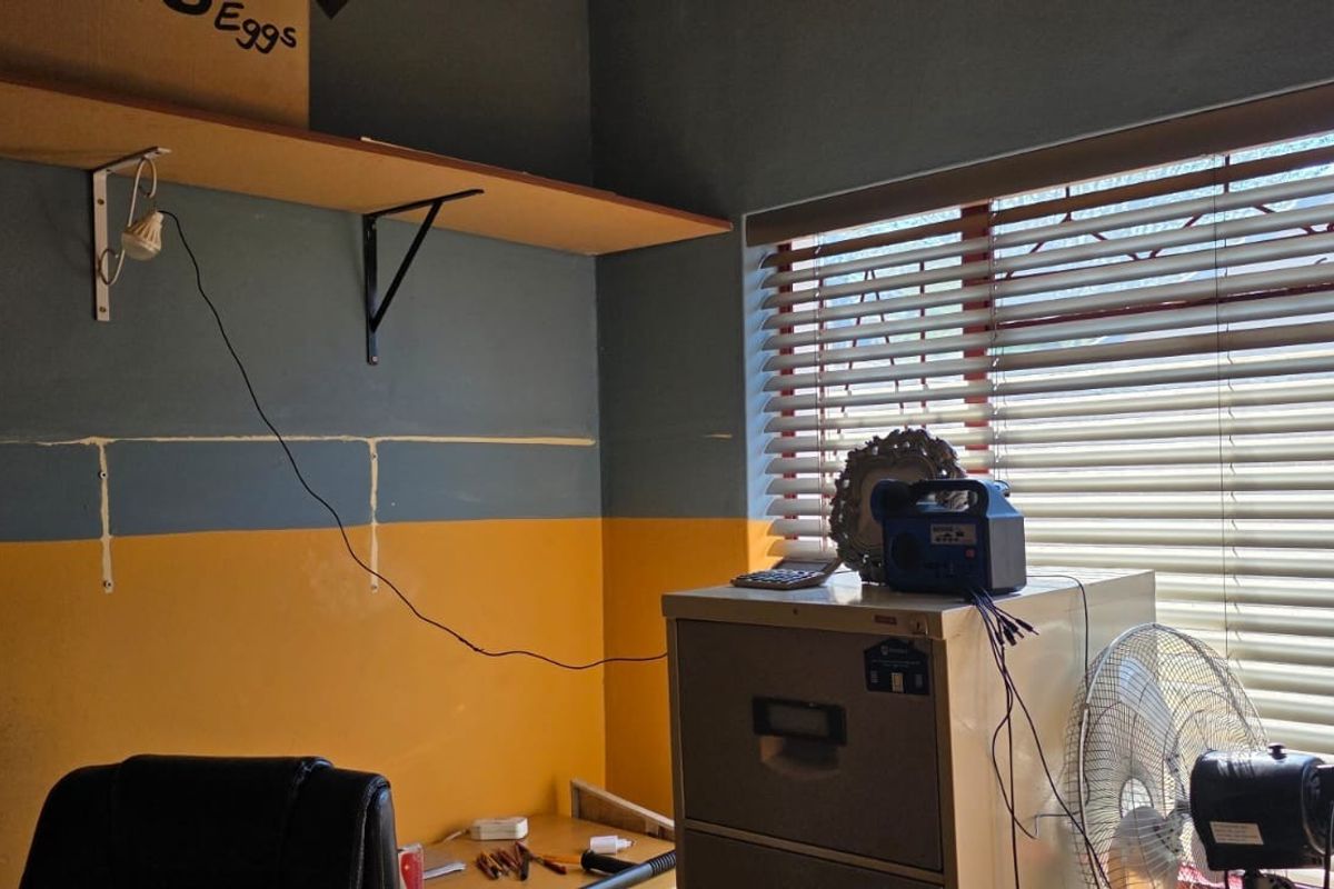 A bedroom space featuring wall-mounted shelving and venetian blinds, currently used as a home office area with visible wall paint inconsistencies.