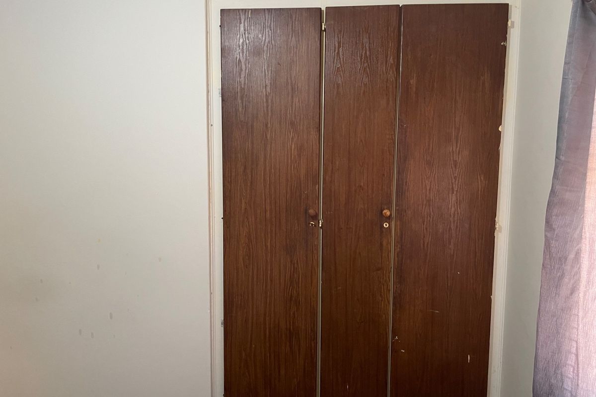 A bedroom interior featuring a dated built-in wooden wardrobe with visible wear and scuffed wall finishes.