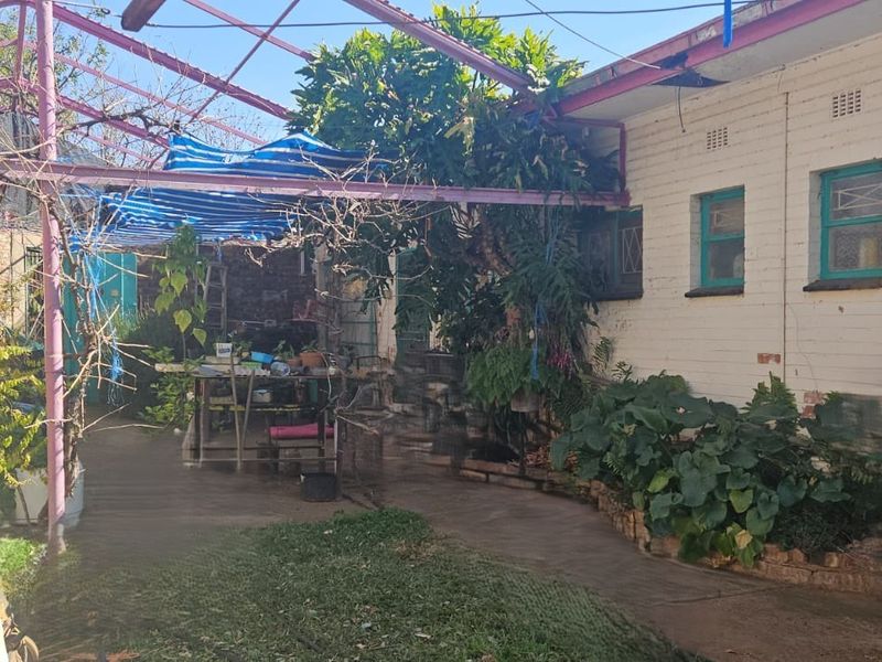 Rear exterior area featuring a metal pergola frame, paved walkway, and brick wall facade, requiring maintenance and landscaping updates.
