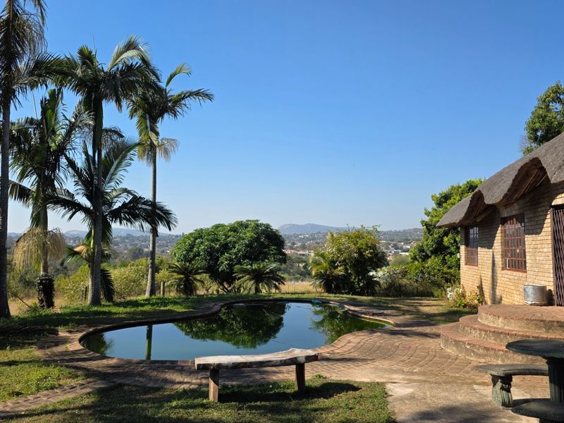 Outdoor pool area featuring a swimming pool, paved patio, and a brick building with a traditional thatched roof, set against a scenic landscape view.