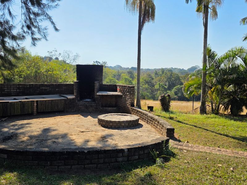 Outdoor entertainment area featuring a built-in brick braai and circular fire pit, set within a landscaped garden with scenic rural views.
