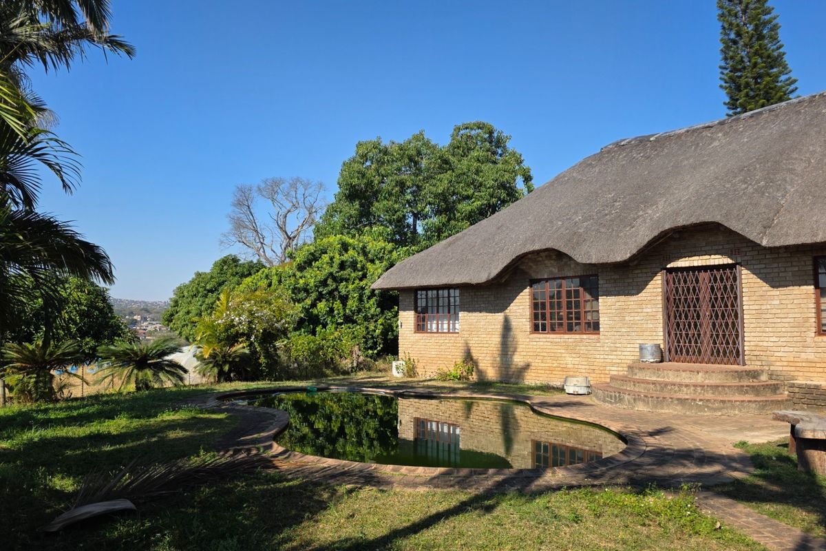 Rear view of a brick farmhouse featuring a thatched roof, a swimming pool, and a surrounding garden area.