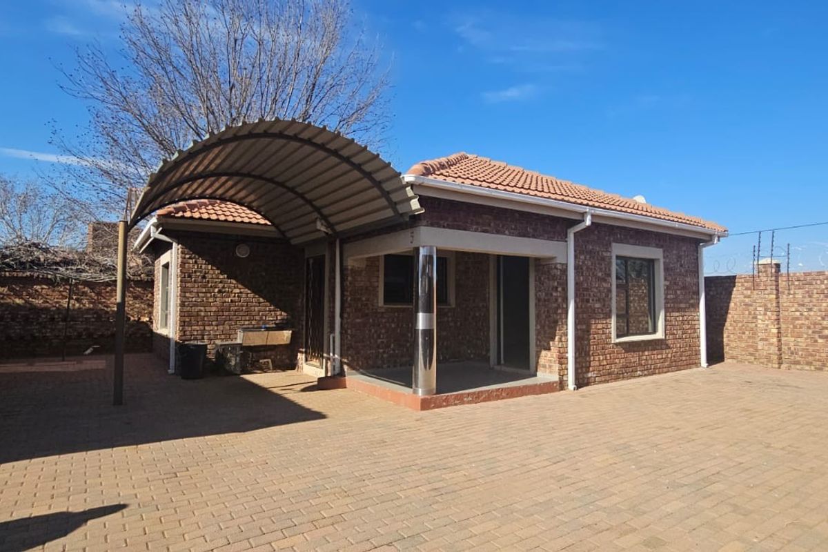 Rear exterior view of a face brick townhouse featuring a paved courtyard, a curved metal carport, and secure perimeter walling with electric fencing.