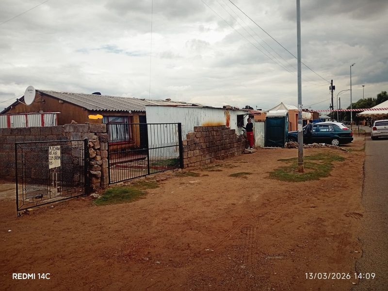 Front view of a residential property featuring a corrugated iron roof, stone boundary wall, and metal gate access from an unpaved street.