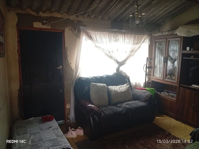 A living room space featuring tiled flooring and an exposed corrugated ceiling, currently requiring significant wall repairs and cosmetic renovation.
