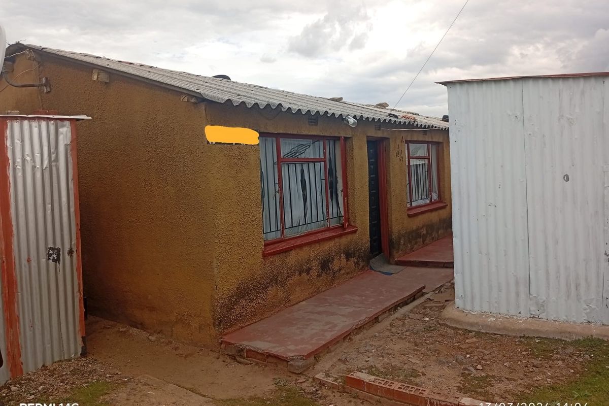 Rear view of a single-story dwelling featuring a corrugated iron roof, security bars on windows, and a concrete pathway.