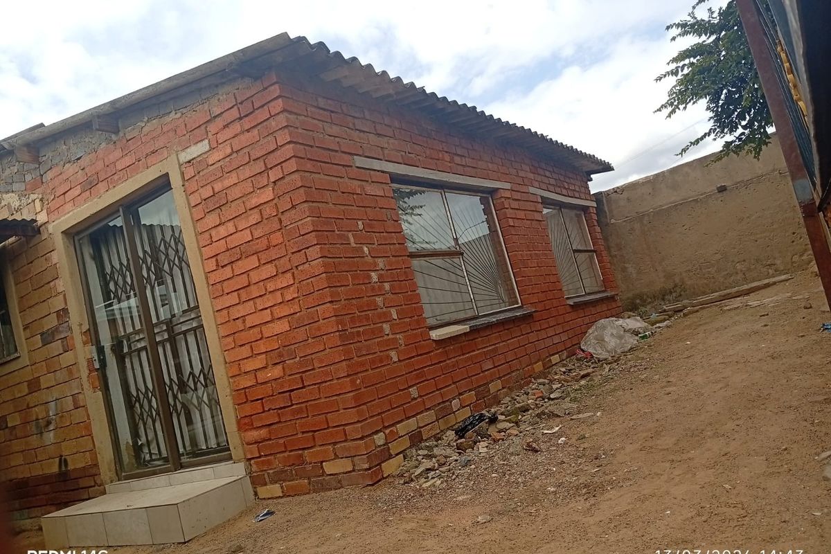 Exterior view of a brick house featuring a sliding glass door with security gate and aluminium window frames, set on an unpaved yard.