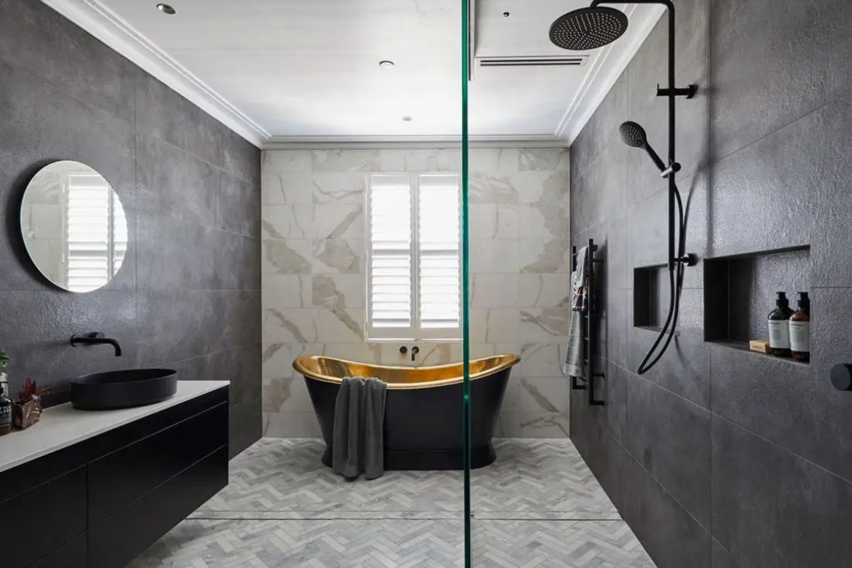 Modern luxury bathroom featuring a gold-accented freestanding bathtub, dark stone wall tiles, herringbone floor tiling, and high-end matte black fixtures.