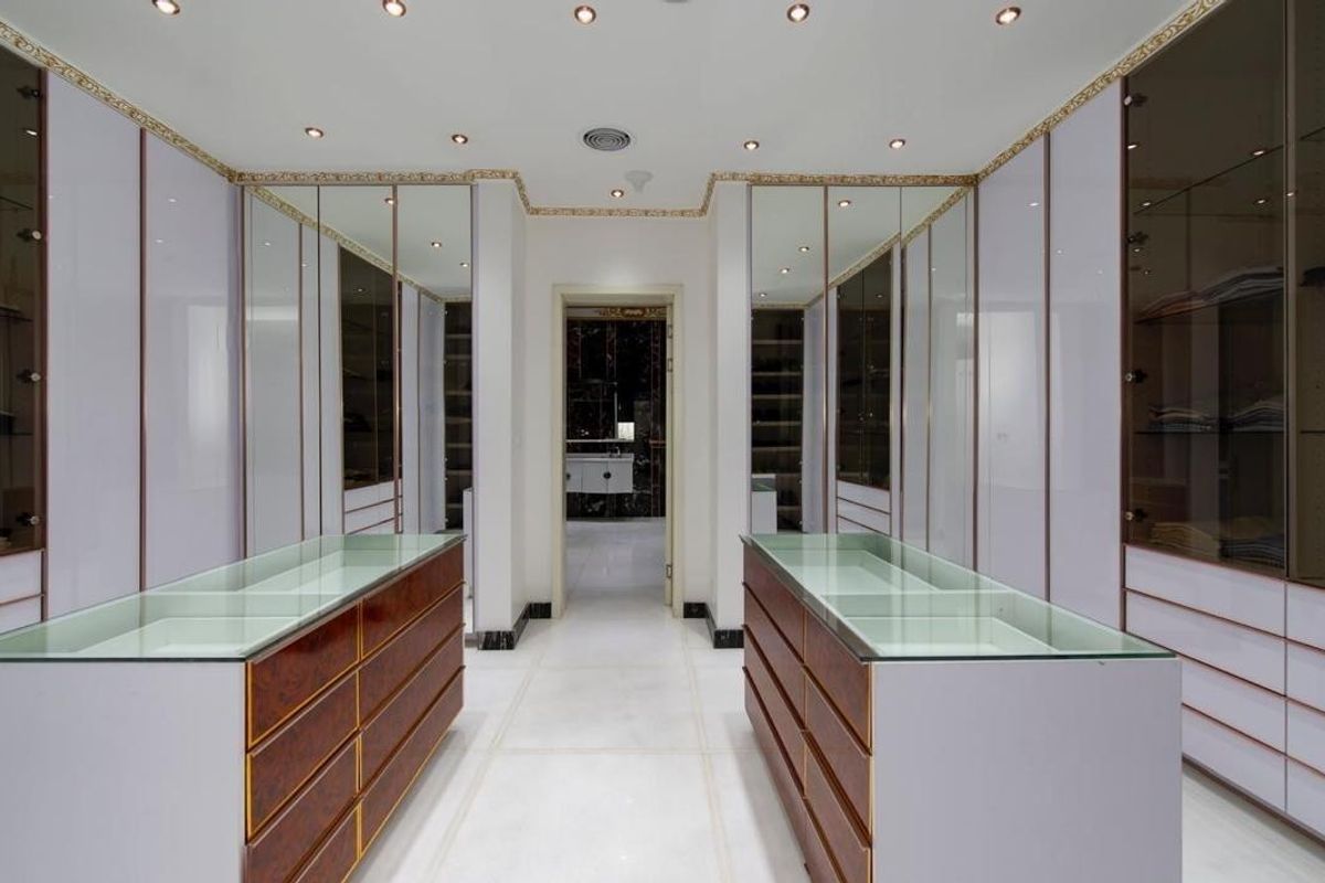 A luxurious walk-in dressing room featuring extensive built-in cabinetry, glass-topped storage islands, marble flooring, and elegant gold-accented crown molding.