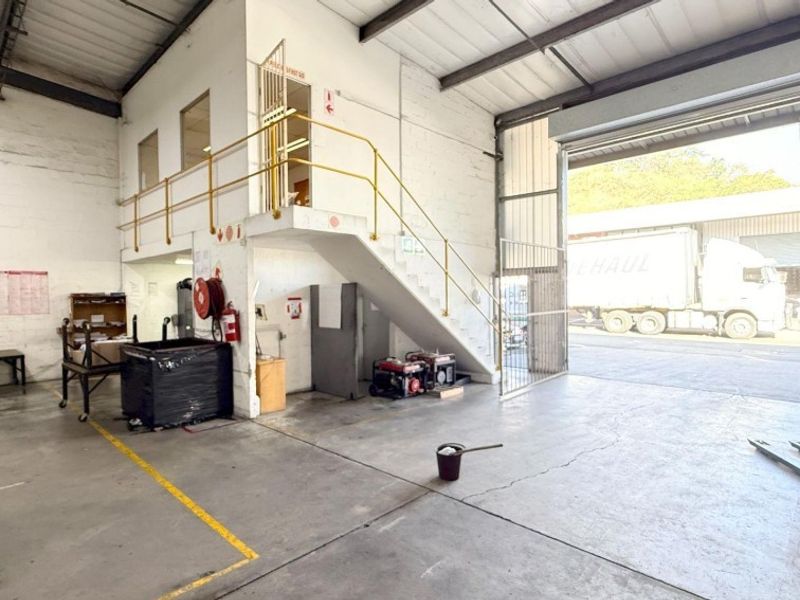 Industrial warehouse space featuring a large roller shutter door, concrete flooring, exposed steel roof trusses, and a mezzanine level with metal staircase access.