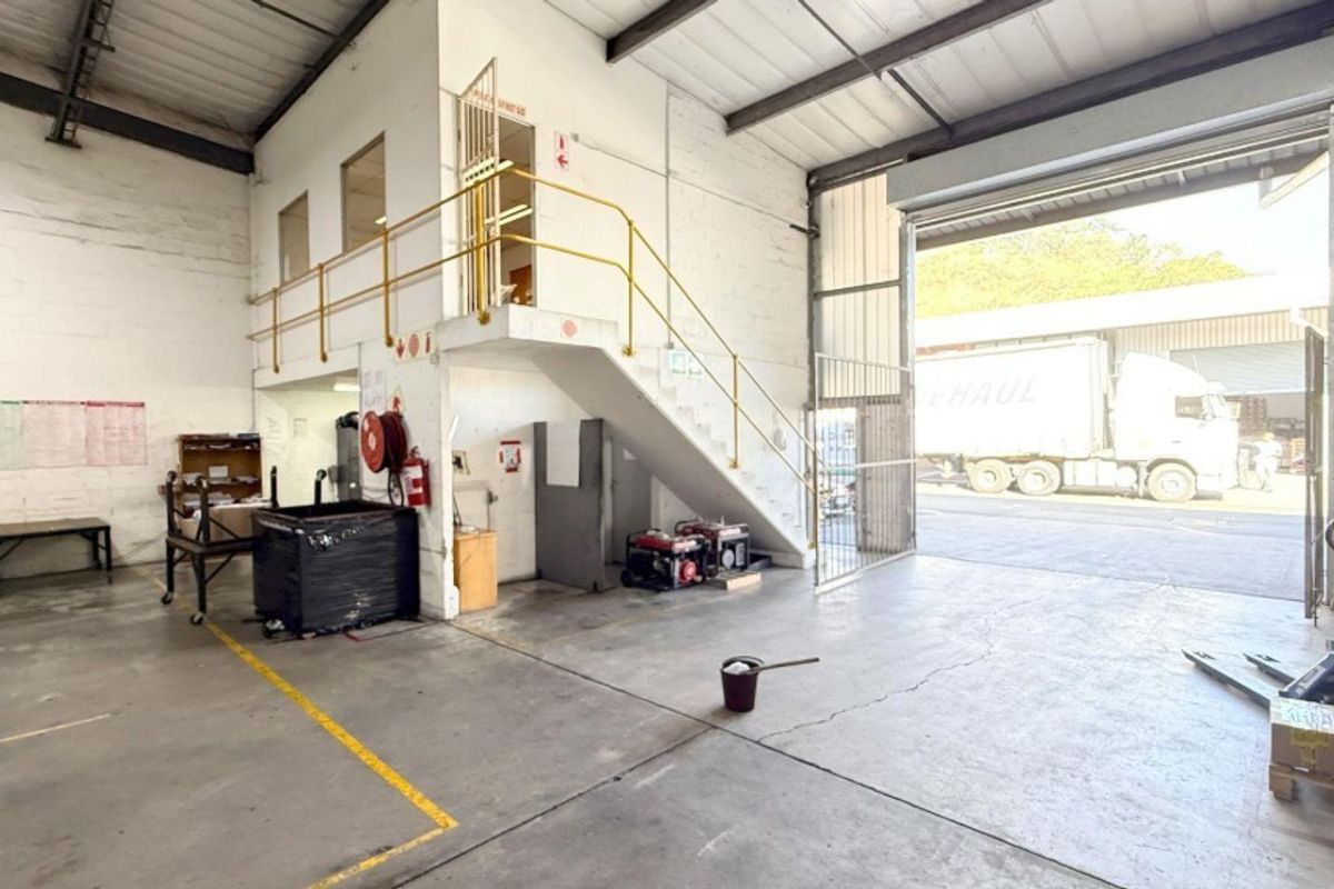 Industrial warehouse space featuring a large roller shutter door, concrete flooring, exposed steel roof trusses, and a mezzanine level with metal staircase access.