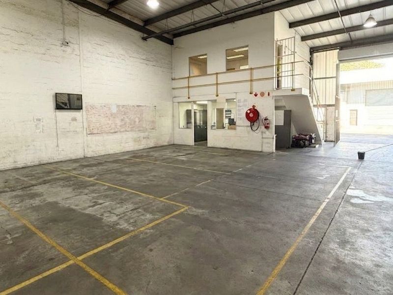 Spacious industrial warehouse unit featuring high ceilings with exposed trusses, a mezzanine level, concrete flooring, and a large roller shutter door for easy access.