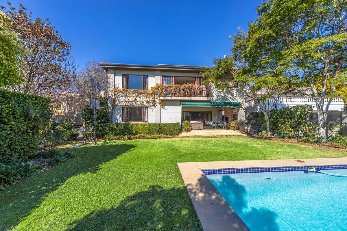 Rear view of a two-story house featuring a swimming pool, landscaped garden, ground-floor patio with retractable awning, and an upper-level balcony.