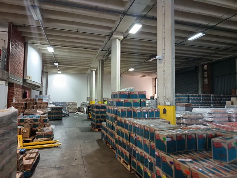 Spacious industrial warehouse area featuring high ceilings with exposed concrete beams, concrete flooring, and functional overhead fluorescent lighting.