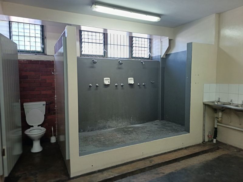 Industrial-style communal bathroom facility featuring multiple shower heads, a flush toilet, and a stainless steel sink with exposed plumbing.