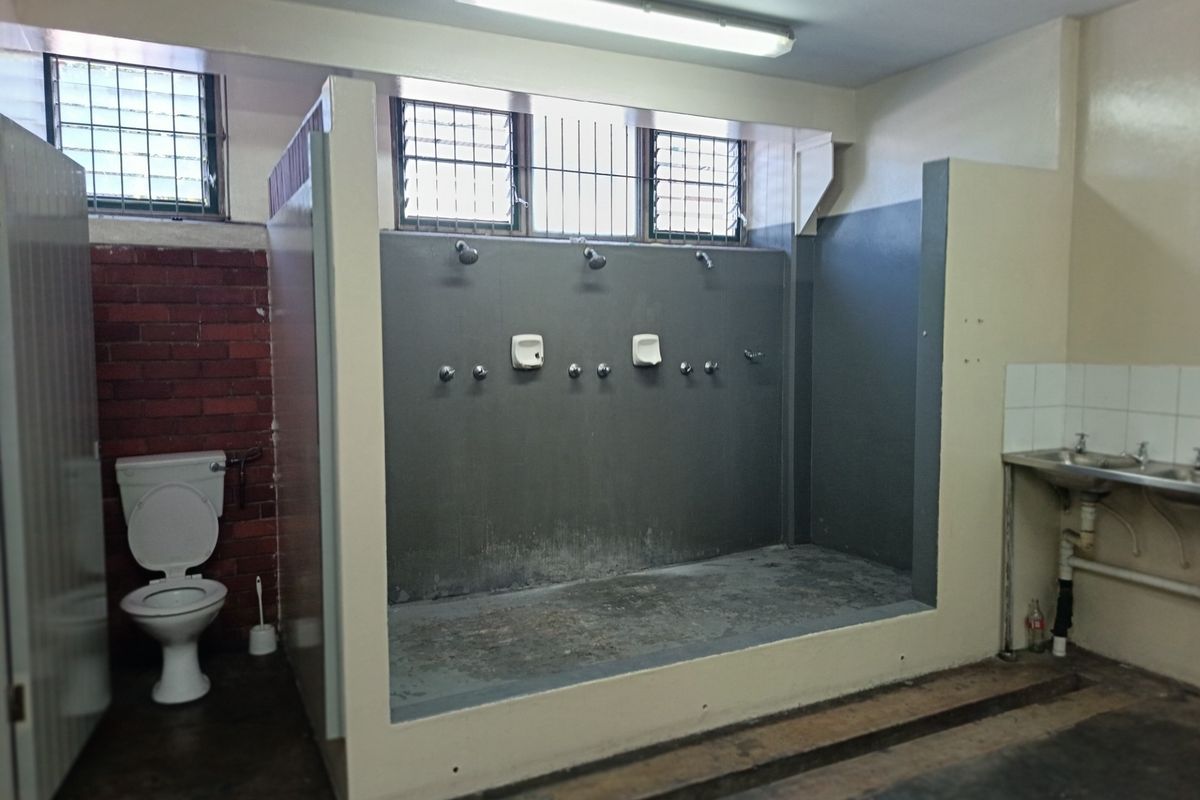 Industrial-style communal bathroom facility featuring multiple shower heads, a flush toilet, and a stainless steel sink with exposed plumbing.