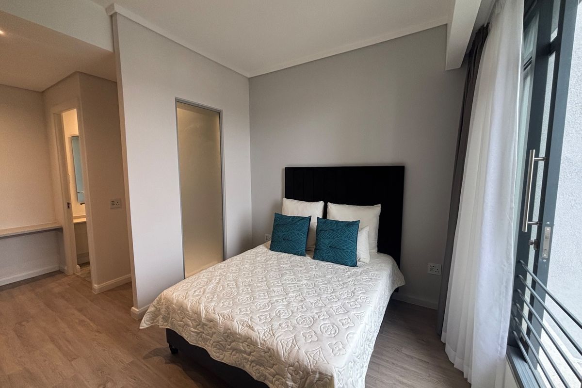 A modern bedroom featuring laminate flooring, neutral walls, and a sliding door leading to a balcony, with an adjacent en-suite bathroom area.