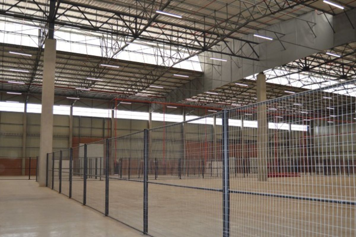 A large, open-plan industrial warehouse space featuring high ceilings with exposed steel trusses, concrete flooring, and secure wire mesh partitioning.