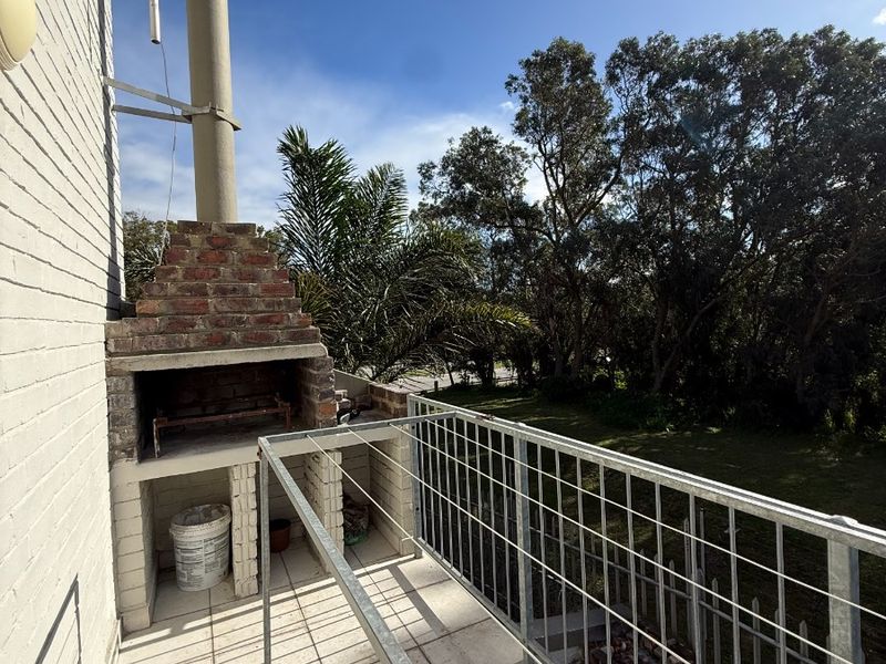 A functional balcony featuring a built-in brick braai, tiled flooring, and a metal safety balustrade overlooking a garden area.