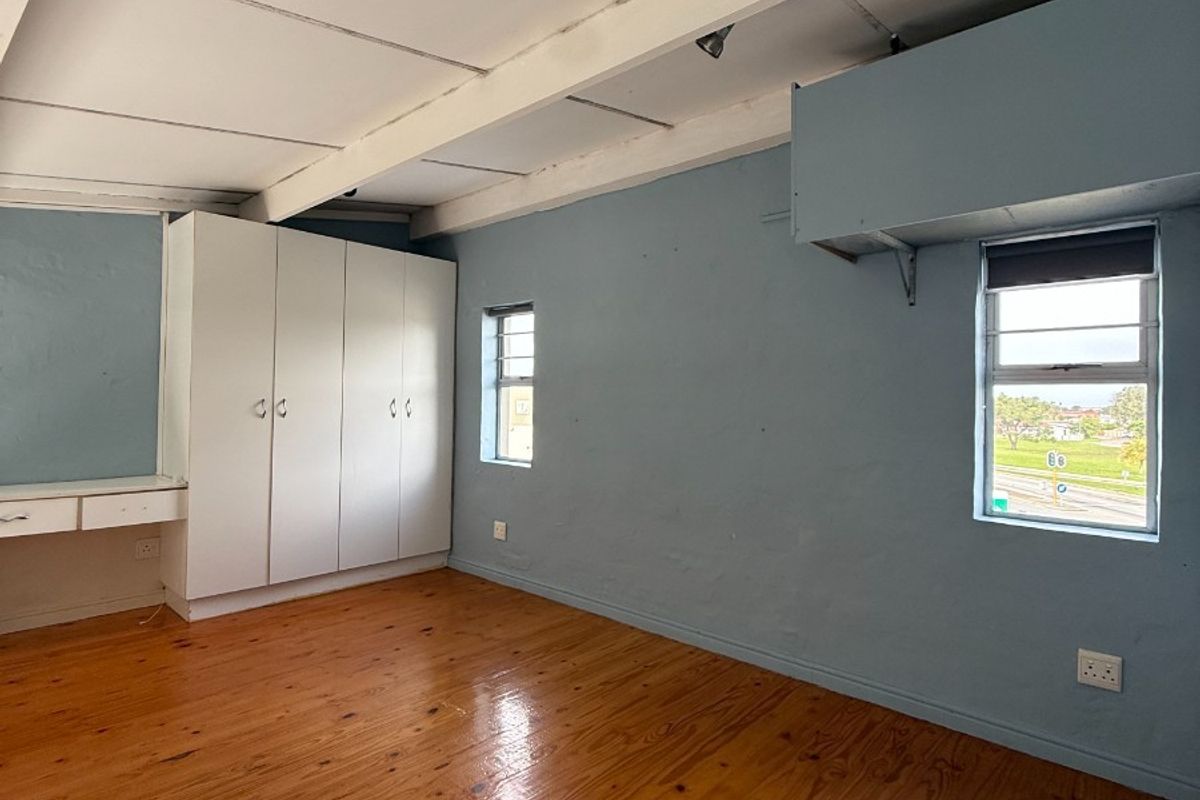 An empty bedroom featuring polished timber flooring, exposed ceiling beams, built-in wardrobe storage, and a integrated desk area with louver windows.