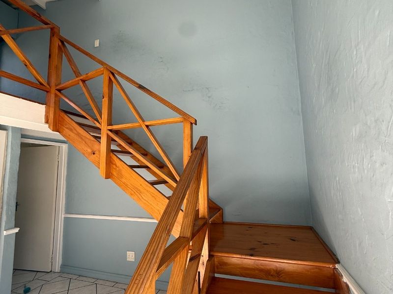 An interior view of a hallway featuring a wooden staircase with a balustrade, tiled flooring, and exposed beam ceiling.